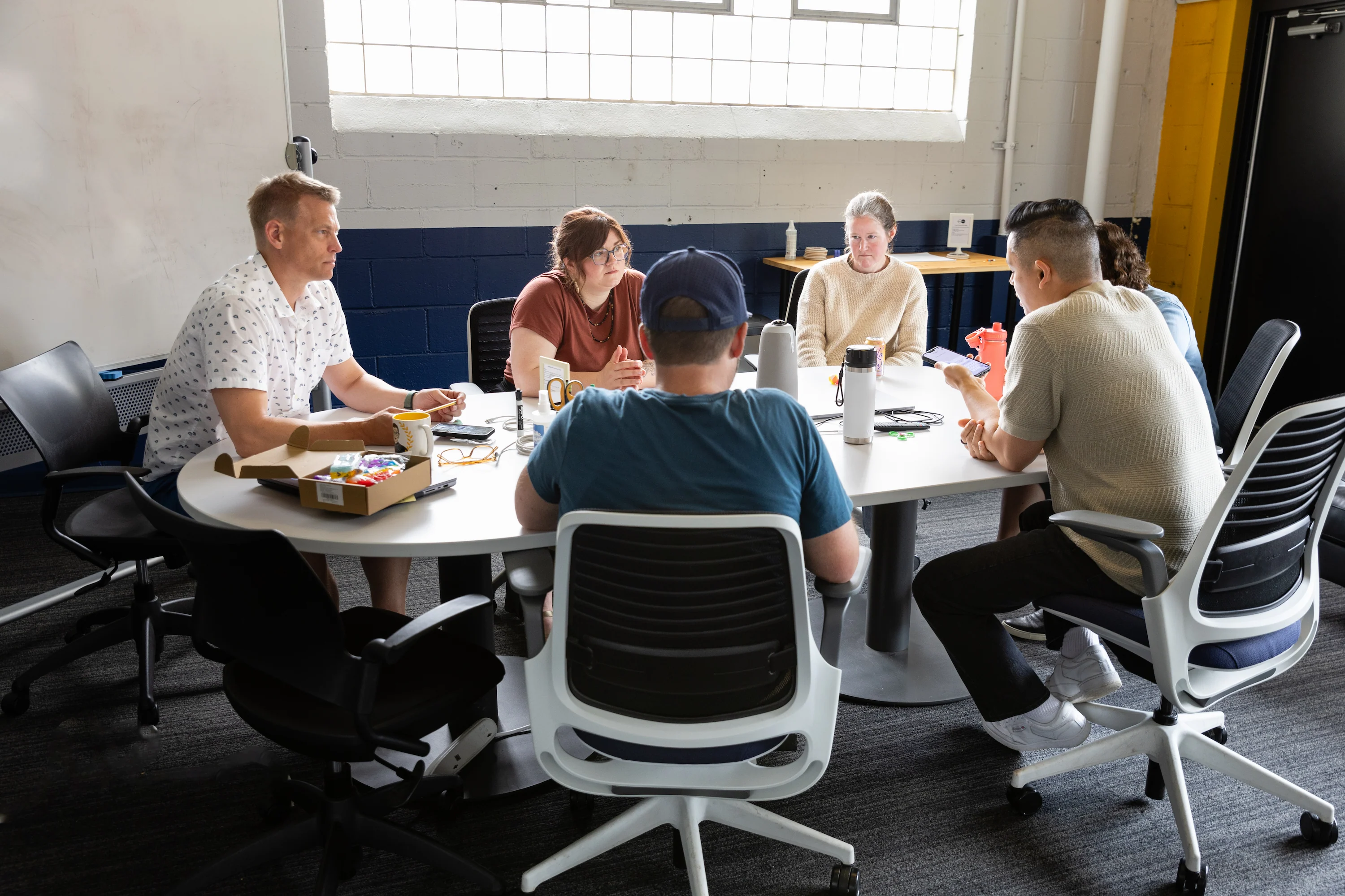 Five people sitting around a round white table in an office setting, engaged in a discussion, with one person showing something on a smartphone while others listen attentively.