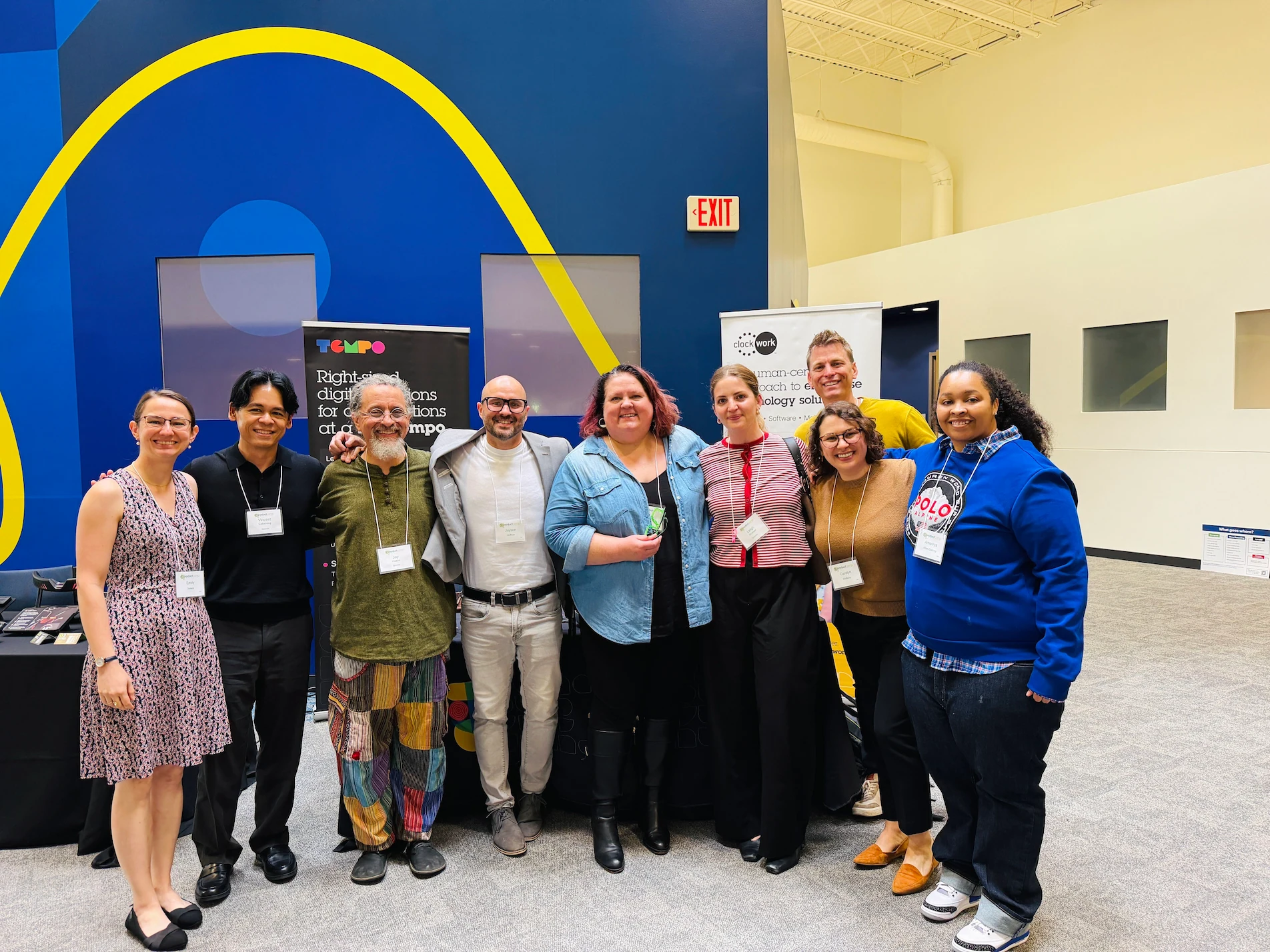 Group of nine people standing indoors in front of two banner stands with text, smiling and posing for a photo. The background includes a blue wall with a yellow curved line, an EXIT sign, and a beige wall with square windows.
