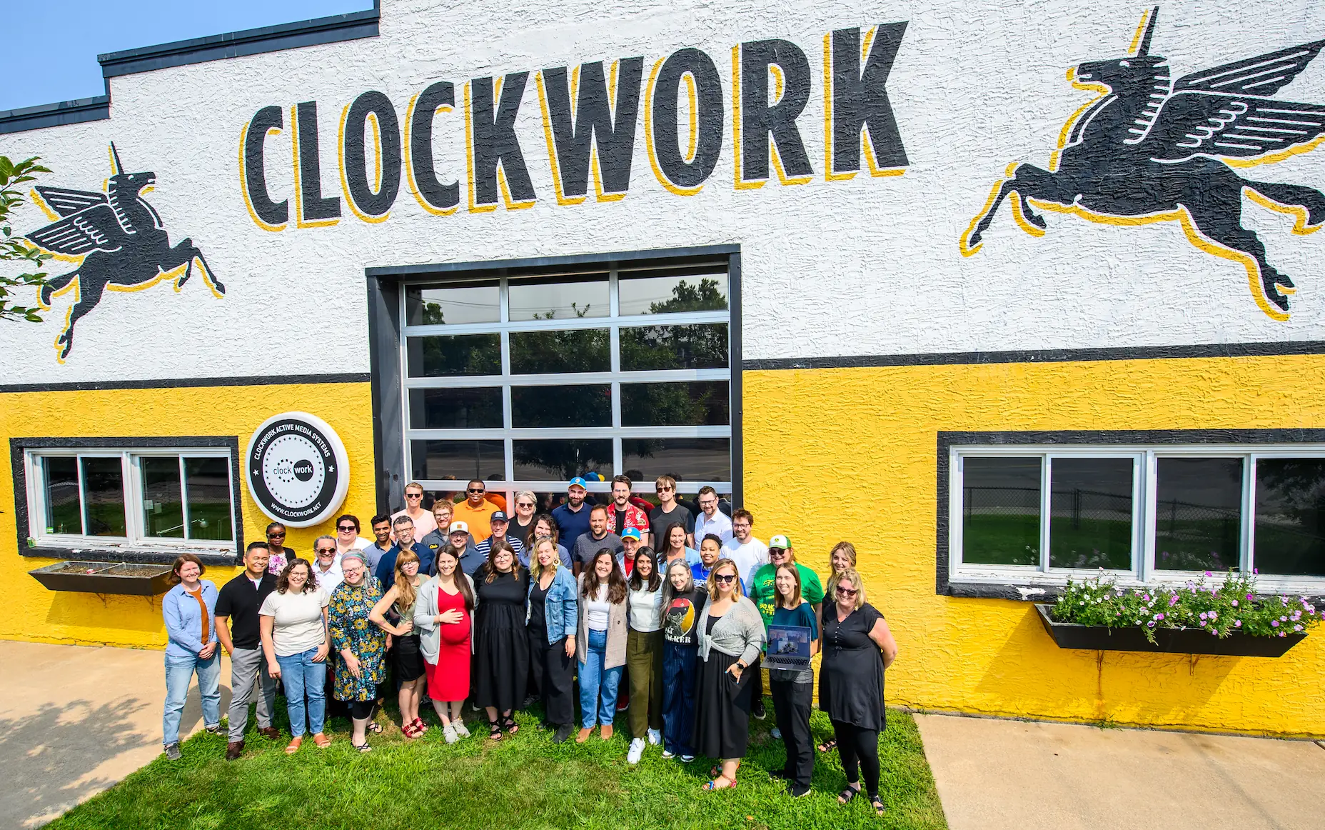 Group of diverse adults standing outside a building with a yellow and white painted wall, large text reading 'CLOCKWORK', and two black winged unicorn murals.