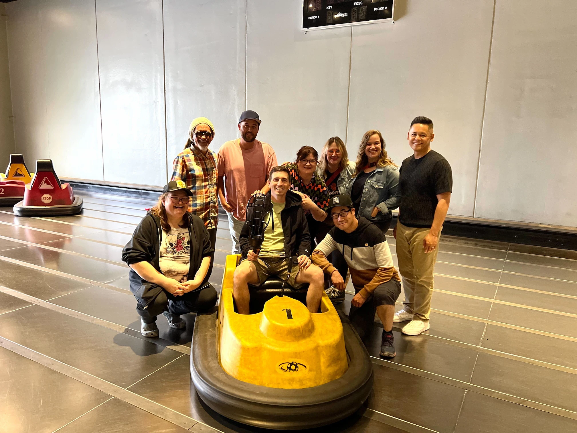 Group of eight people posing together on an indoor bumper car track; one person sits in a yellow bumper car marked with the number 1 and a black steering wheel, while the others stand or crouch around them, smiling. The background shows a plain wall and parts of other bumper cars.
