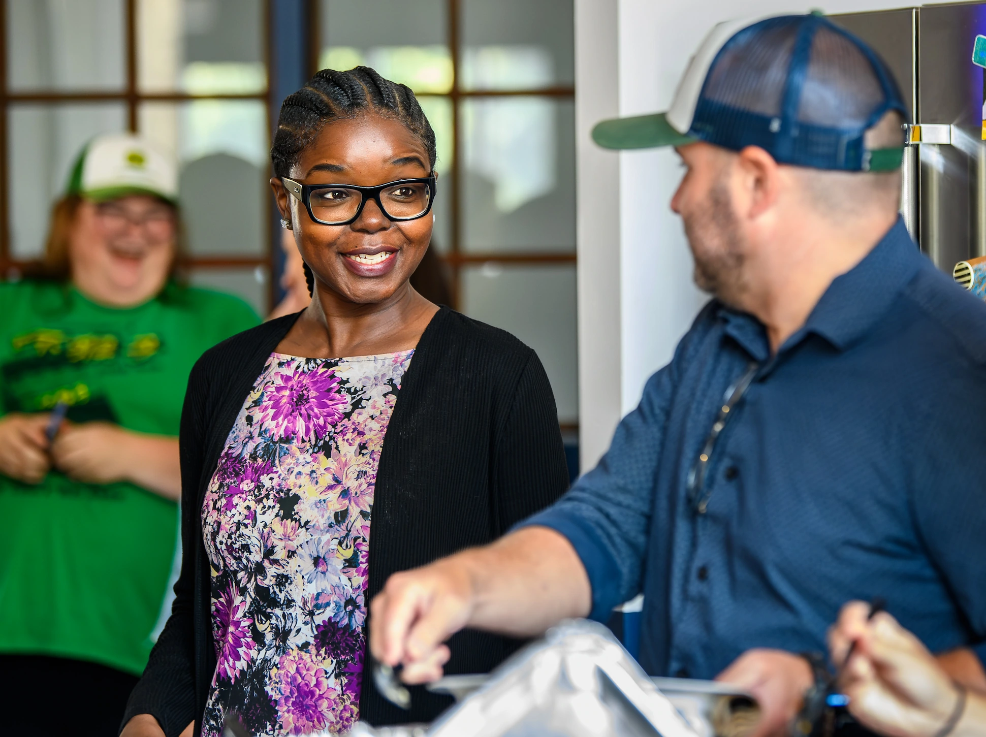 Smiling woman with glasses and braided hair wearing a floral top and black cardigan, talking to a man in a blue shirt and cap, with another person laughing in the background.