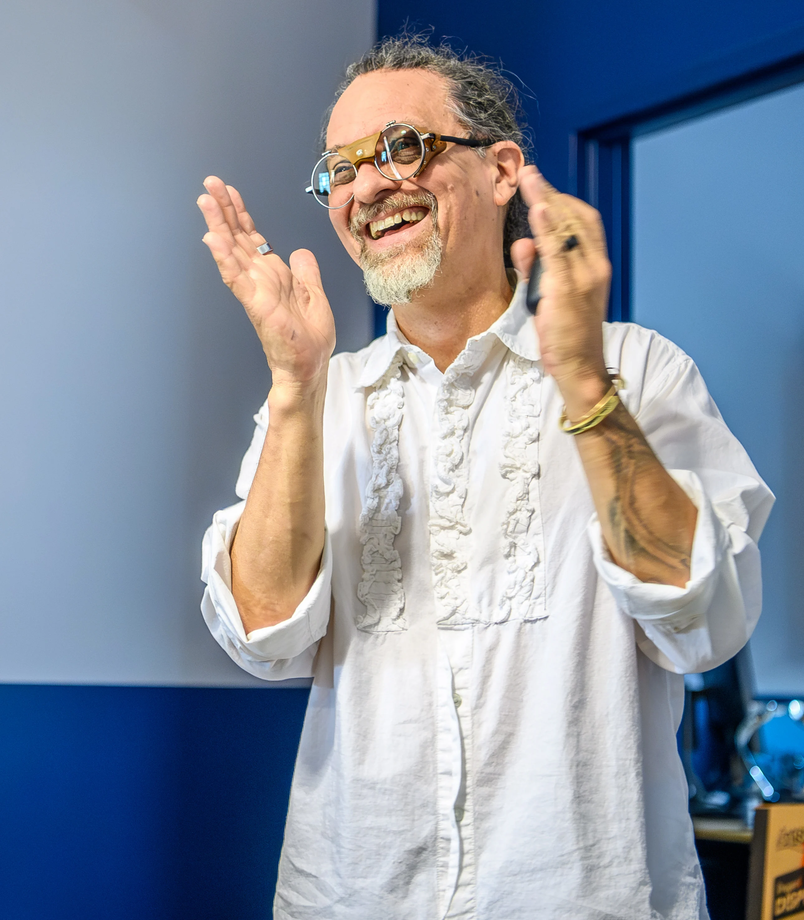 Smiling man with glasses clapping his hands, wearing a white embroidered shirt, standing in a room with blue and gray walls.