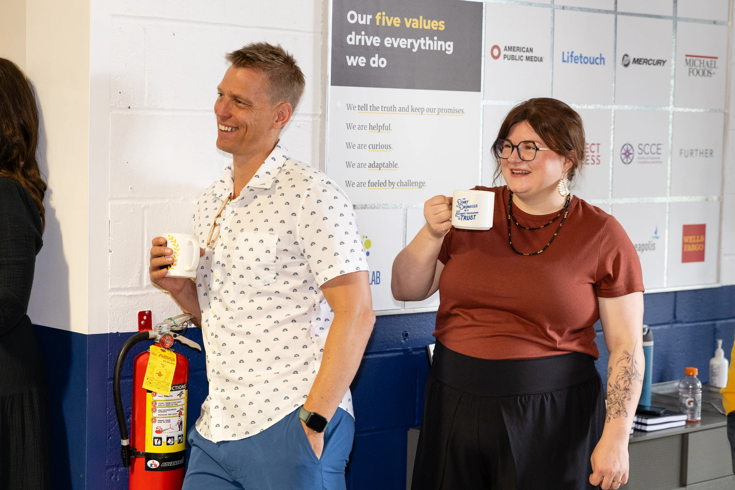 Two people standing and smiling in an office setting, each holding a white coffee mug. A man in a white patterned short-sleeve shirt and blue pants leans against a wall with a fire extinguisher, while a woman with glasses, wearing a rust-colored top and black skirt, stands nearby. Behind them is a wall with a poster displaying 'Our five values drive everything we do' and logos of various organizations.