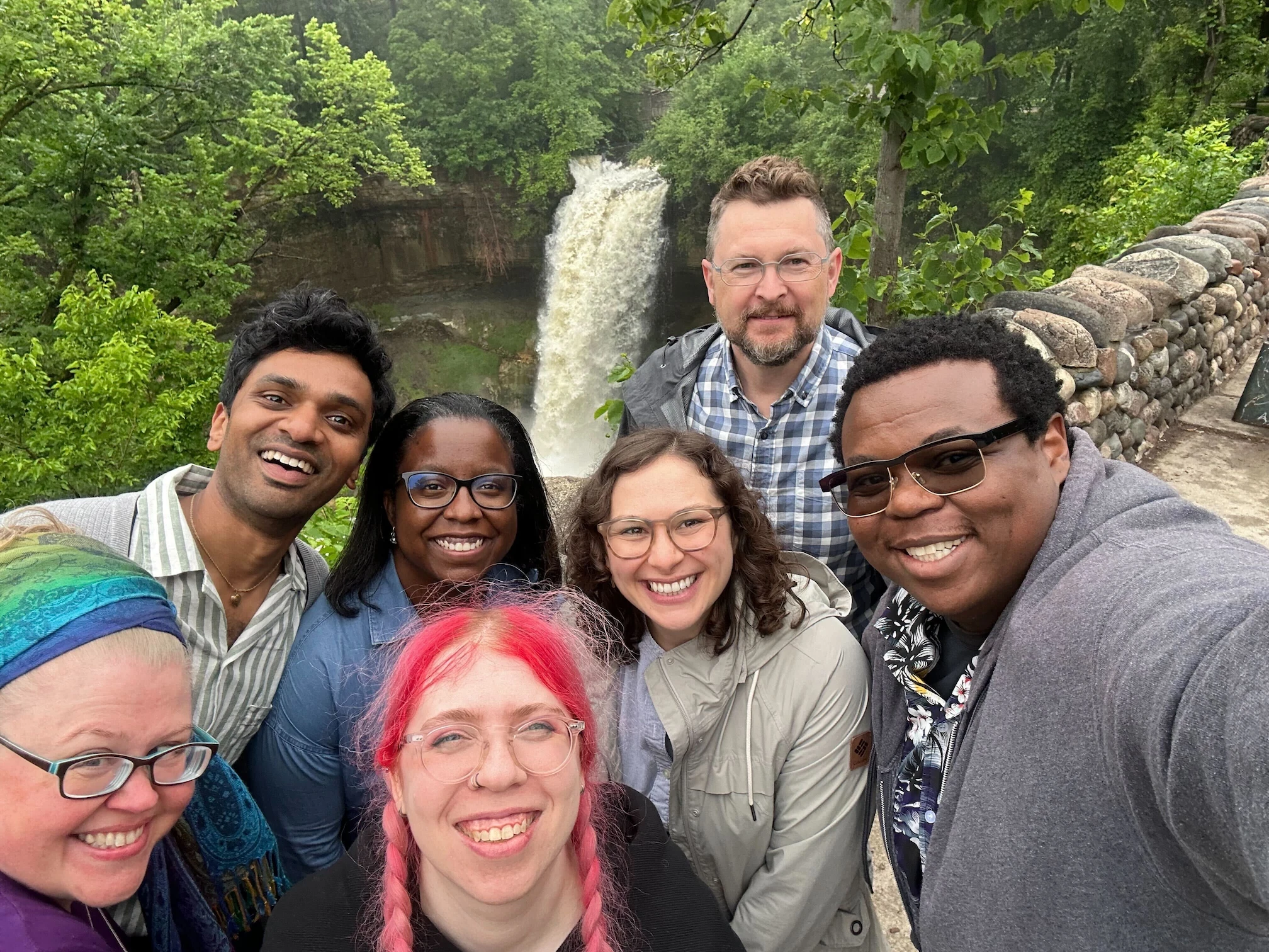 Diverse group of seven smiling adults taking a selfie outdoors in front of a waterfall surrounded by green trees and a stone wall.