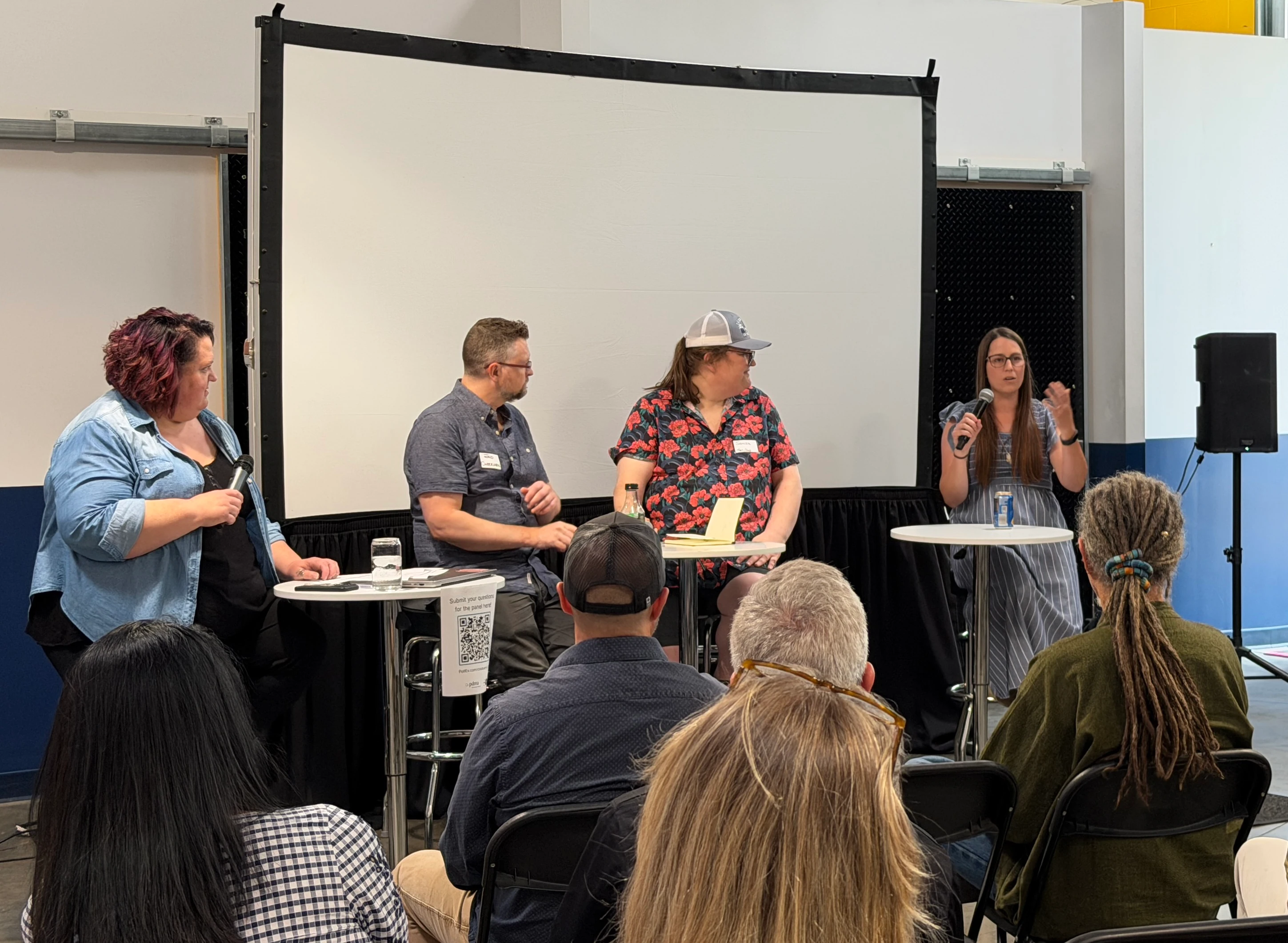 Panel discussion with four people seated on stools in front of a large white screen, two holding microphones, and an audience of six visible from behind watching attentively.