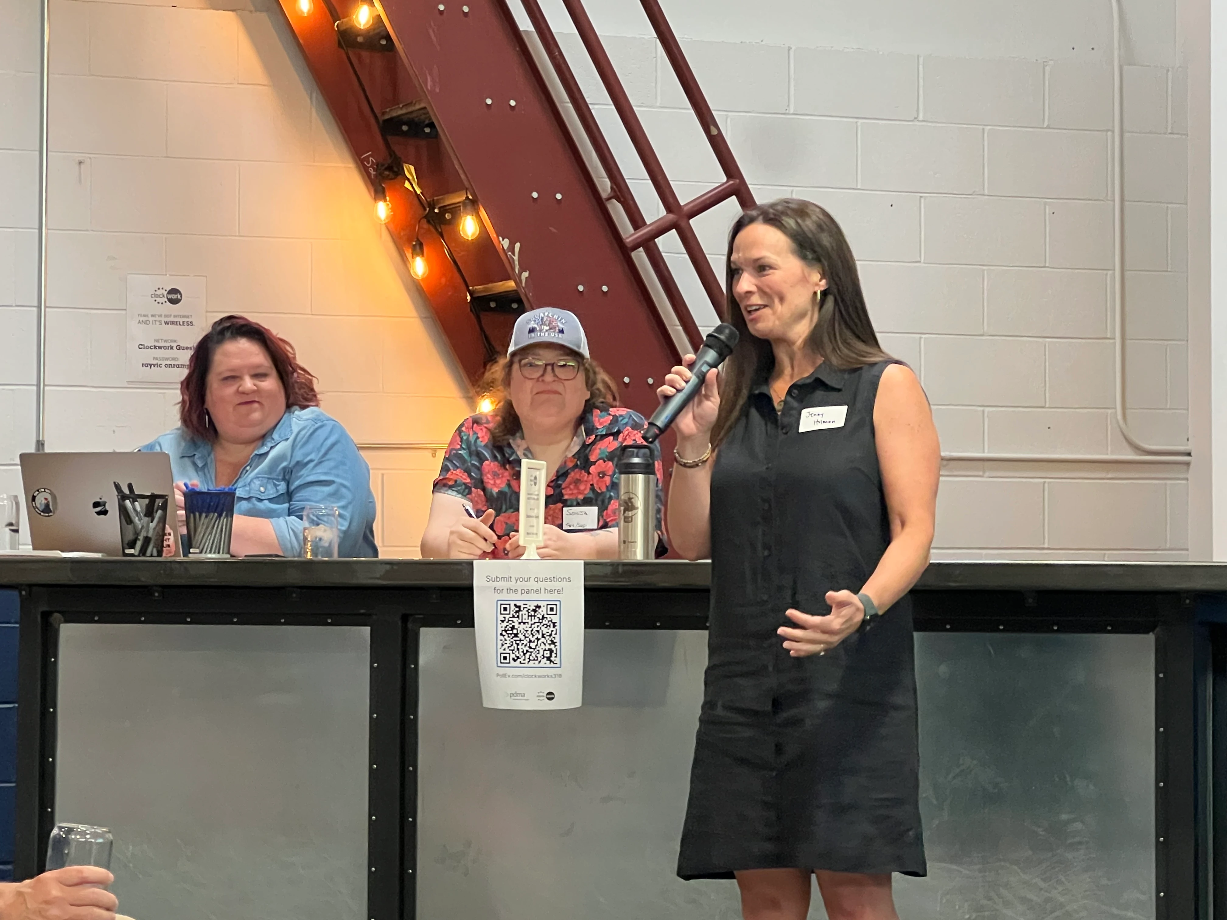 Three women at a panel event: one standing and speaking into a microphone wearing a black sleeveless dress with a name tag, two seated behind a counter with pens, laptop, and drinks; a lit staircase is behind them.