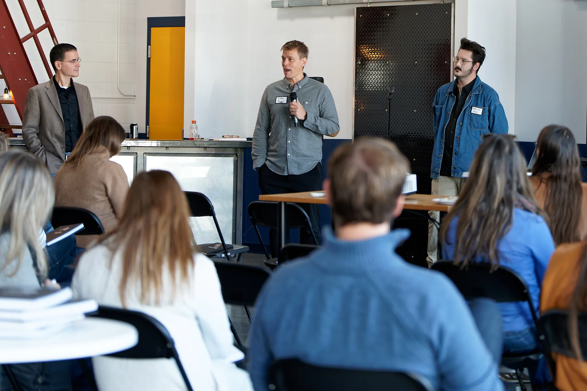 A man in a gray shirt holding a microphone speaks to an audience seated on black chairs, while two other men stand attentively nearby in a room with industrial decor.
