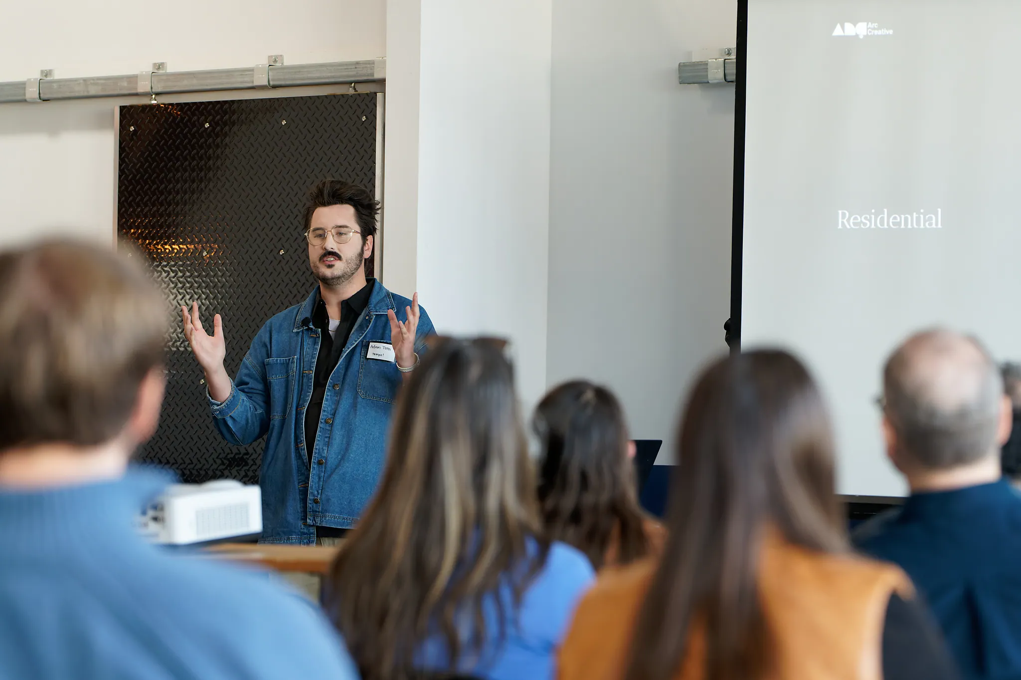Man with glasses and a denim jacket giving a presentation to an audience, with a projection screen showing the word 'Residential'.