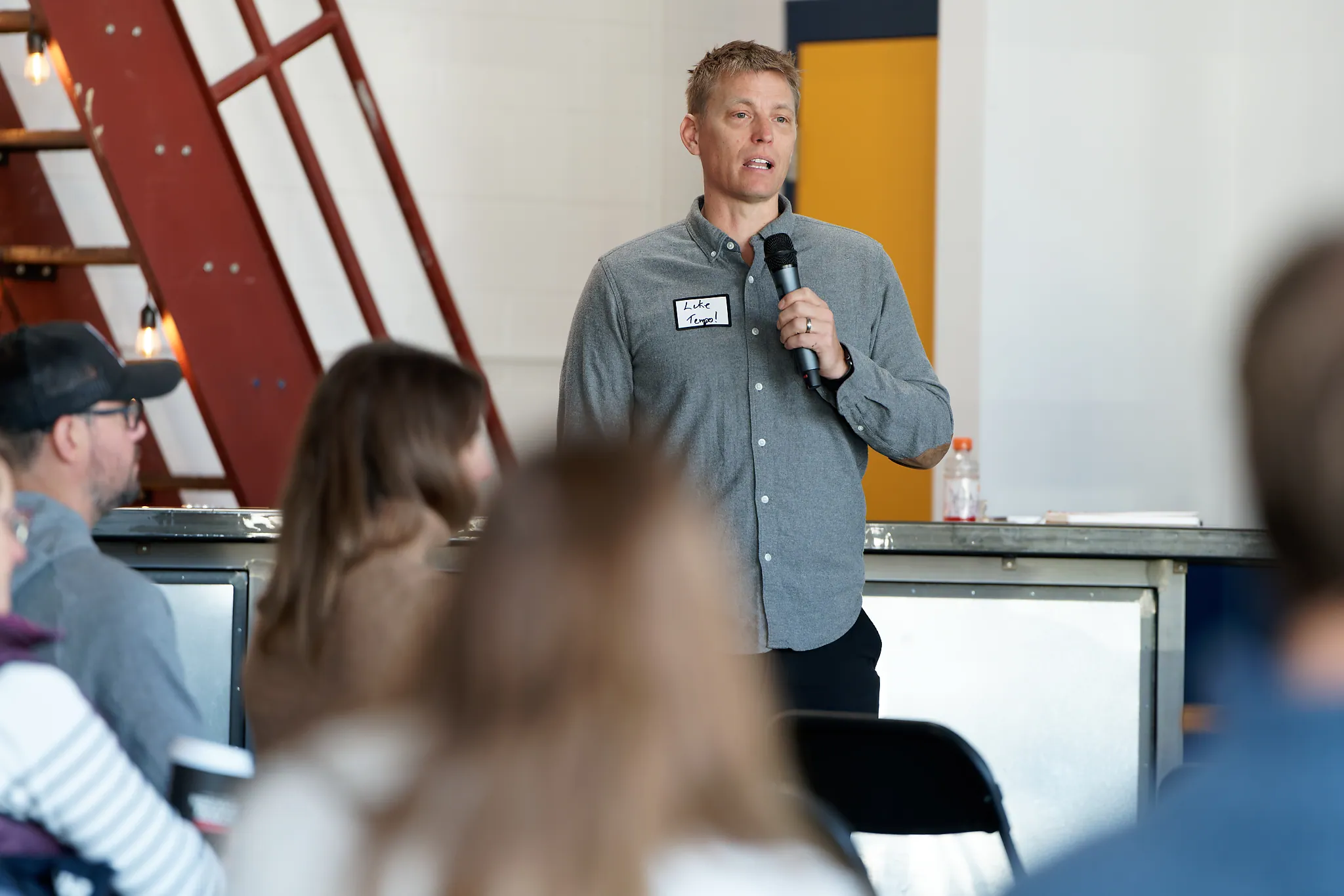 Man in a gray button-up shirt holding a microphone and speaking to an audience in an indoor setting with a red metal stair structure in the background.