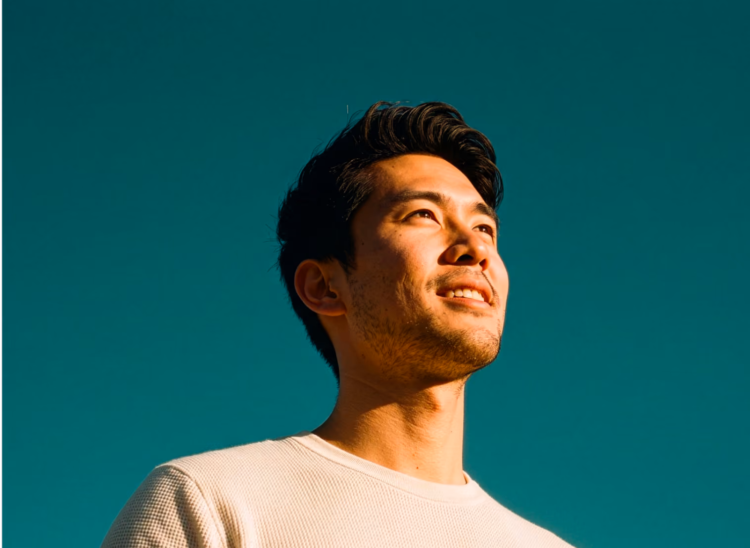 Close-up portrait of a smiling young man with dark hair looking upwards against a clear teal background, wearing a light-colored shirt.