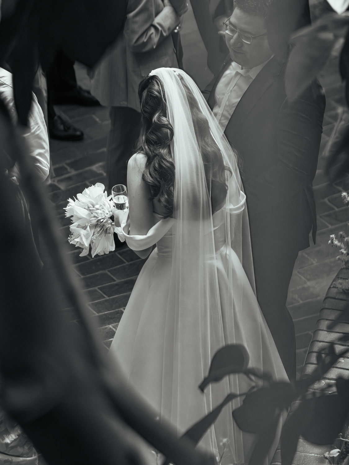 Black and white photo of a bride with long wavy hair wearing a veil and wedding gown, holding a bouquet and a glass, standing next to a smiling groom in a suit, with blurred guests and foliage around.