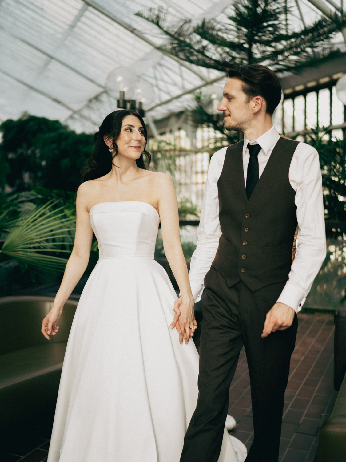 A bride and groom holding hands and looking at each other in a bright greenhouse with plants and glass ceiling, the bride wears a strapless white wedding gown and the groom wears a white shirt, black tie, and dark vest with pants.