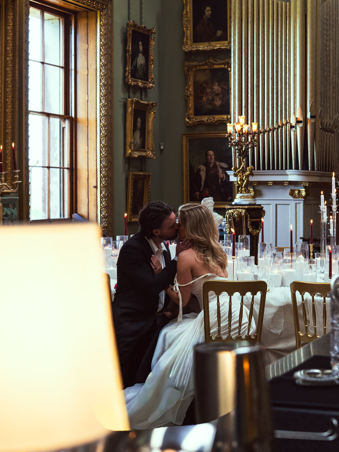 Couple dressed in wedding attire kissing while seated at an elegantly decorated table with lit candles in a richly furnished room featuring large windows, classical paintings in ornate frames, and a large pipe organ.