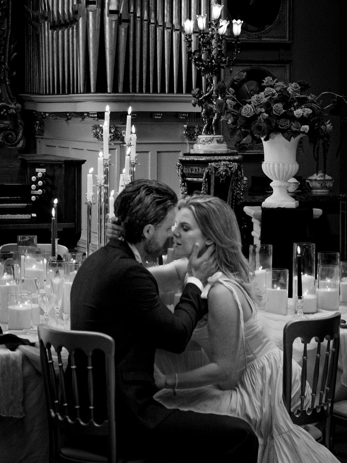 Black and white photo of a couple sitting close on chairs at a candlelit table, leaning in for an intimate moment in an ornately decorated room with flowers, candles, and a large pipe organ in the background.