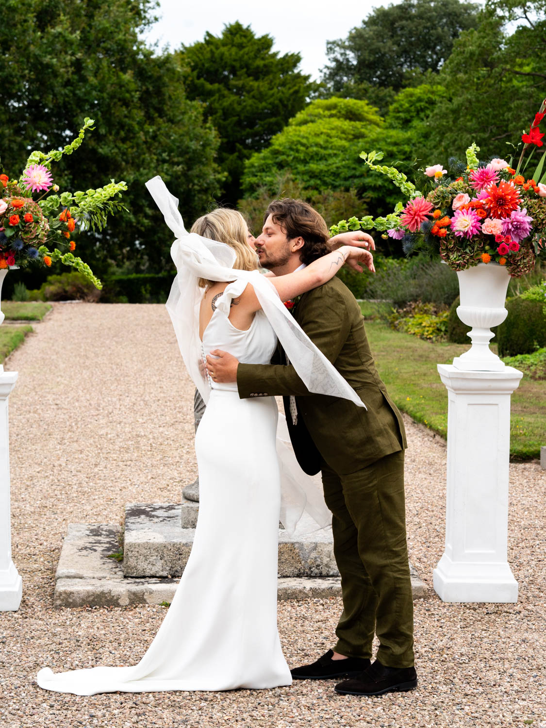 Bride in a white dress and veil embracing and kissing groom in a green suit on a gravel path between two large floral arrangements on white pedestals, with lush green trees and bushes in the background.