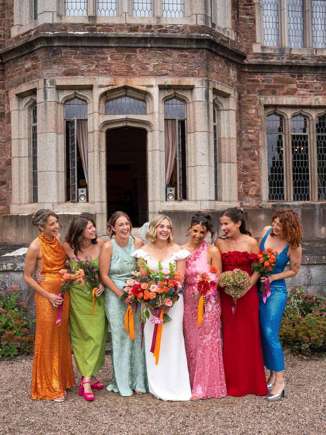 Seven women standing arm in arm outside a stone building with large windows, dressed in colorful formal gowns holding bouquets, smiling and laughing, the woman in the center wearing a white bridal dress and veil.