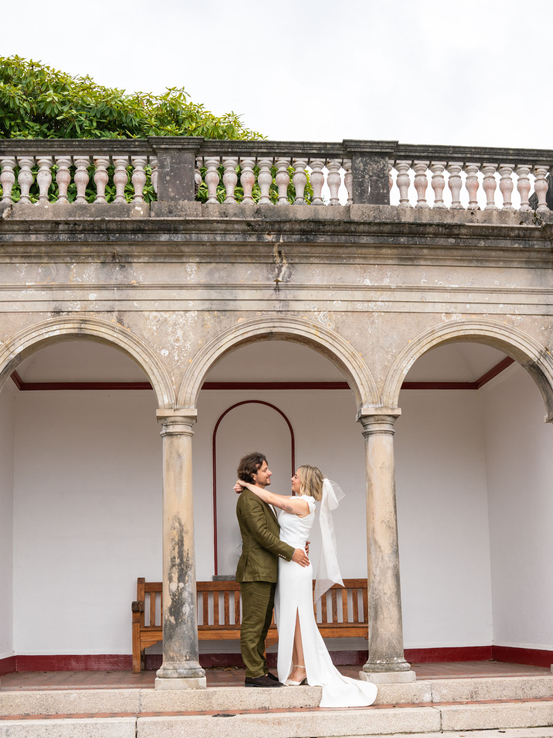 Bride in white dress and veil embracing groom in green suit standing between stone arches with wooden bench behind them.