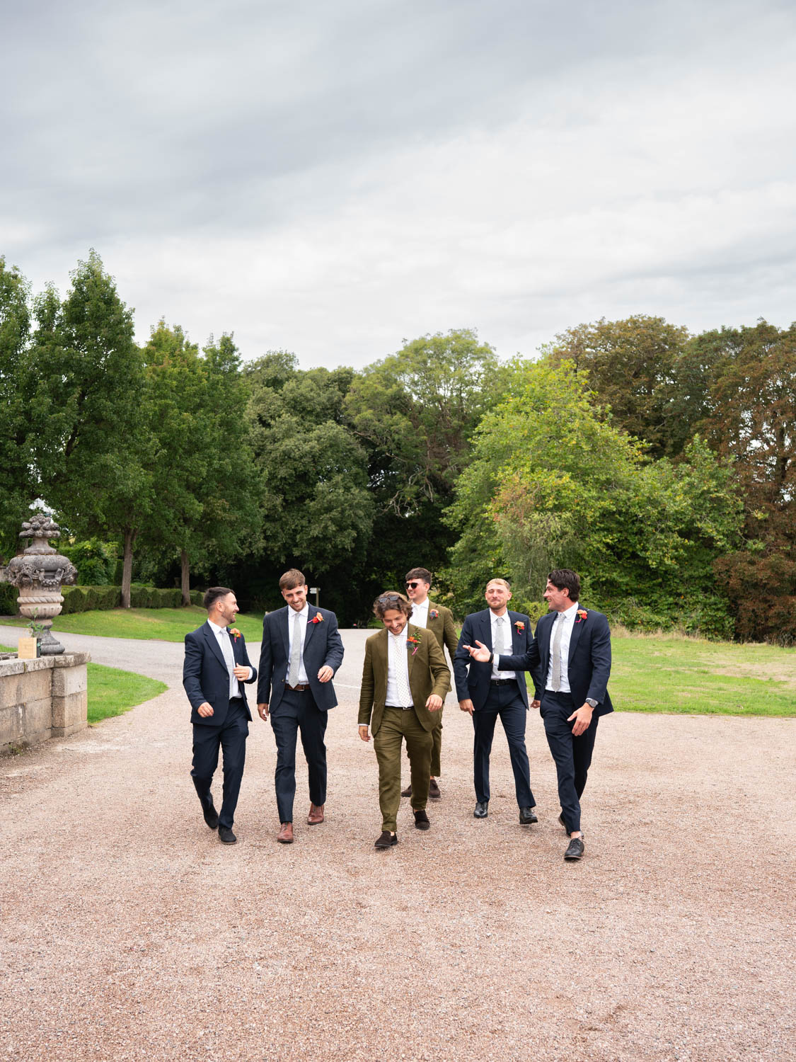 Six well-dressed men walking on a gravel path outdoors with green trees and grass in the background, some wearing navy blue suits and one in a brown suit, all with boutonnieres.