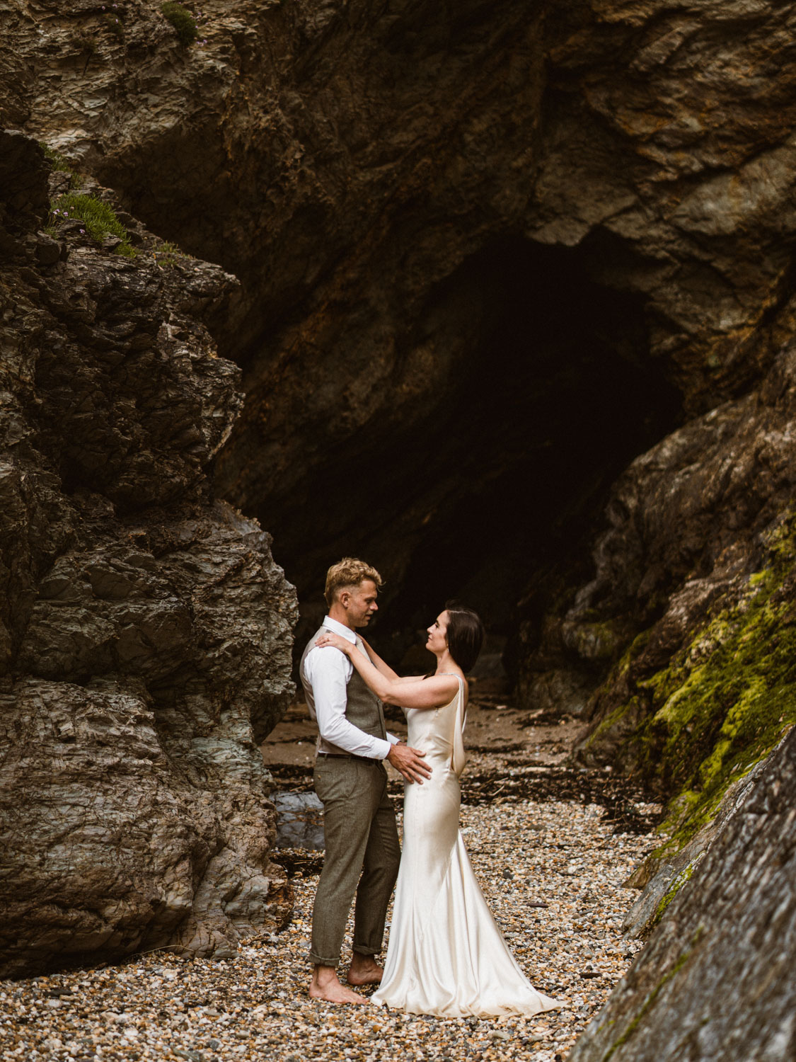 A barefoot couple dressed in wedding attire stands facing each other on a rocky beach near a dark cave entrance; the bride wears a satin white gown and the groom wears a vest and rolled-up pants.