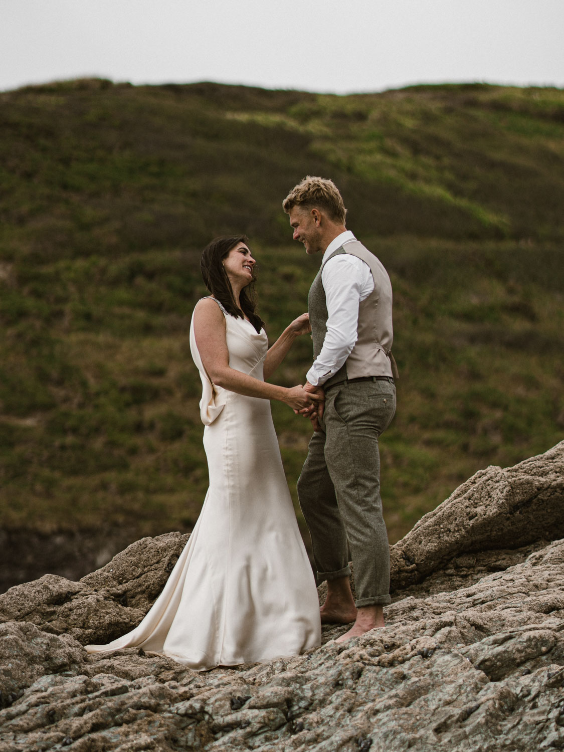 Bride and groom holding hands and looking at each other while standing barefoot on rocky terrain with grassy hills in the background.
