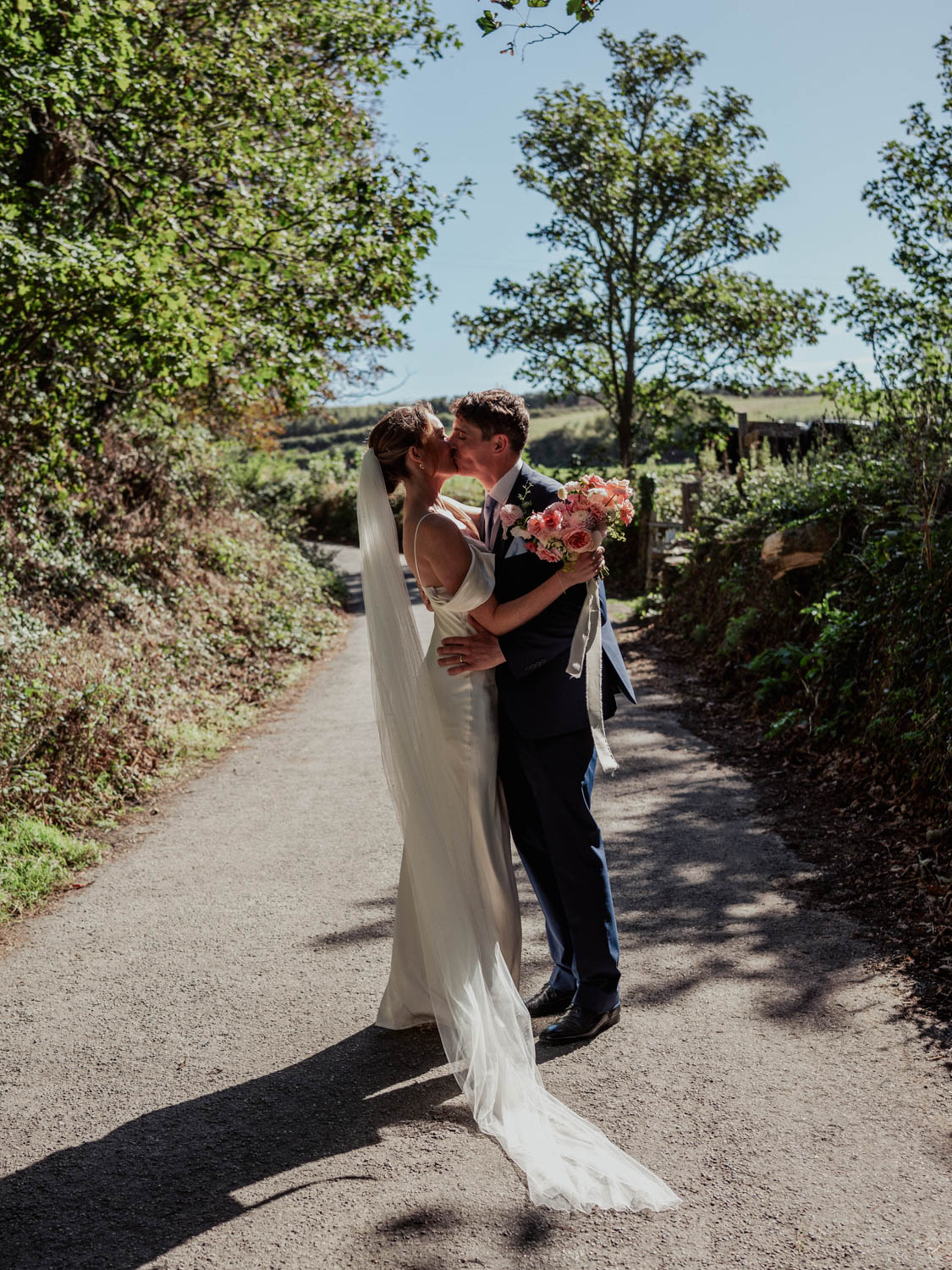 Bride and groom kissing outdoors on a sunny day, standing on a paved path surrounded by greenery; the bride wears a long white gown with a veil and holds a bouquet of pink flowers, the groom wears a dark suit.