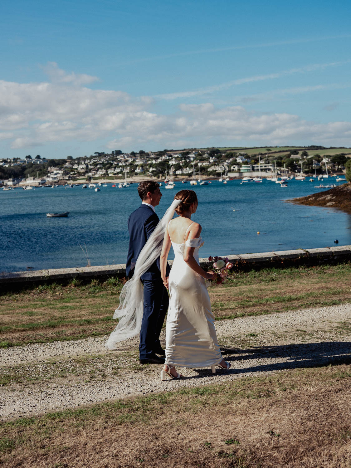A bride and groom walk hand in hand on a gravel path beside a body of water with boats and a village visible across the water under a partly cloudy blue sky.