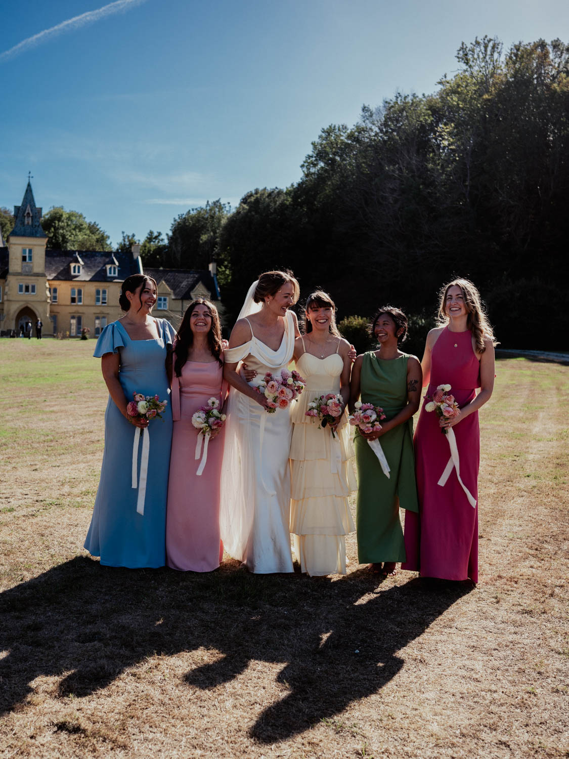 Bride in white wedding dress with veil standing outside on dry grass next to five bridesmaids wearing long dresses in blue, pink, cream, green, and magenta, all holding bouquets and smiling with a large yellow house and trees in the background under a bright blue sky.