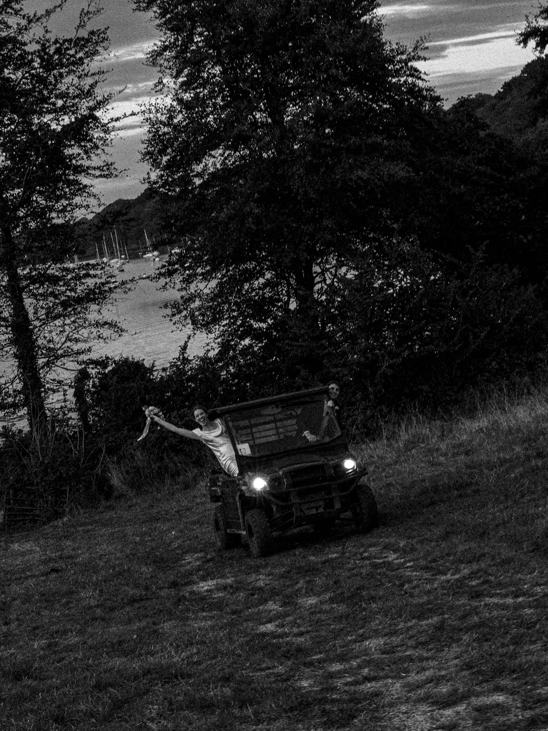 Black-and-white photo of a couple riding an off-road vehicle down a grassy hill with the woman leaning out of the side holding up a bouquet, and sailboats visible in the water behind trees.