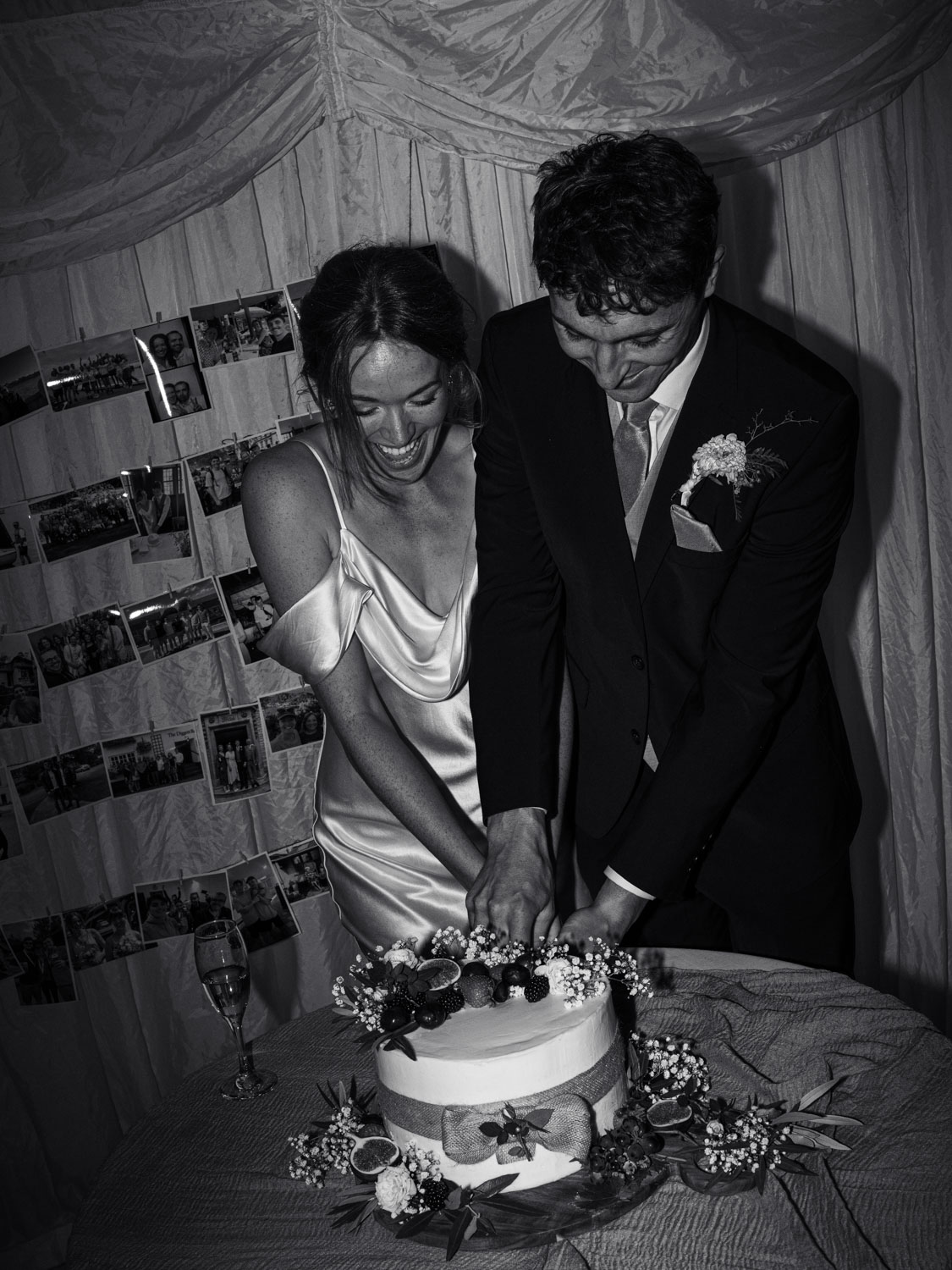 Bride and groom smiling and jointly cutting a decorated two-tier wedding cake at their reception, with photos displayed on the wall behind them.
