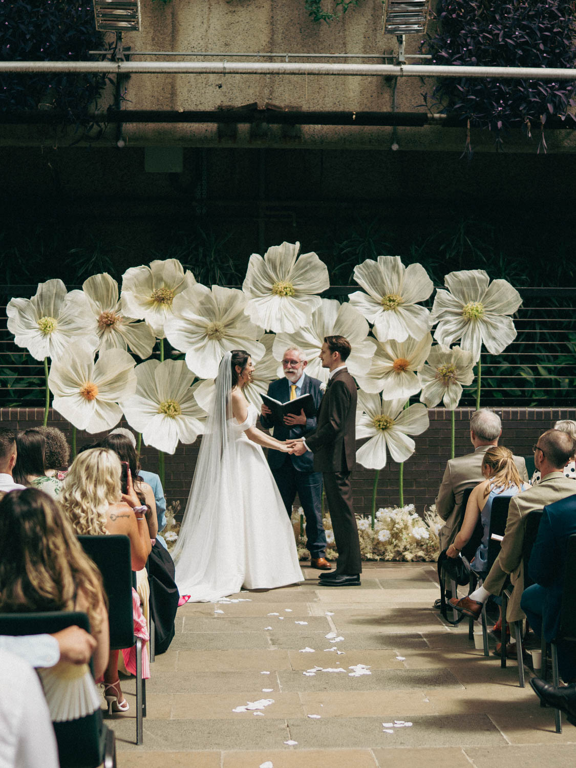 Bride in white gown and veil and groom in dark suit holding hands facing an officiant during an indoor wedding ceremony, with large white flower decorations behind them and guests seated on both sides.