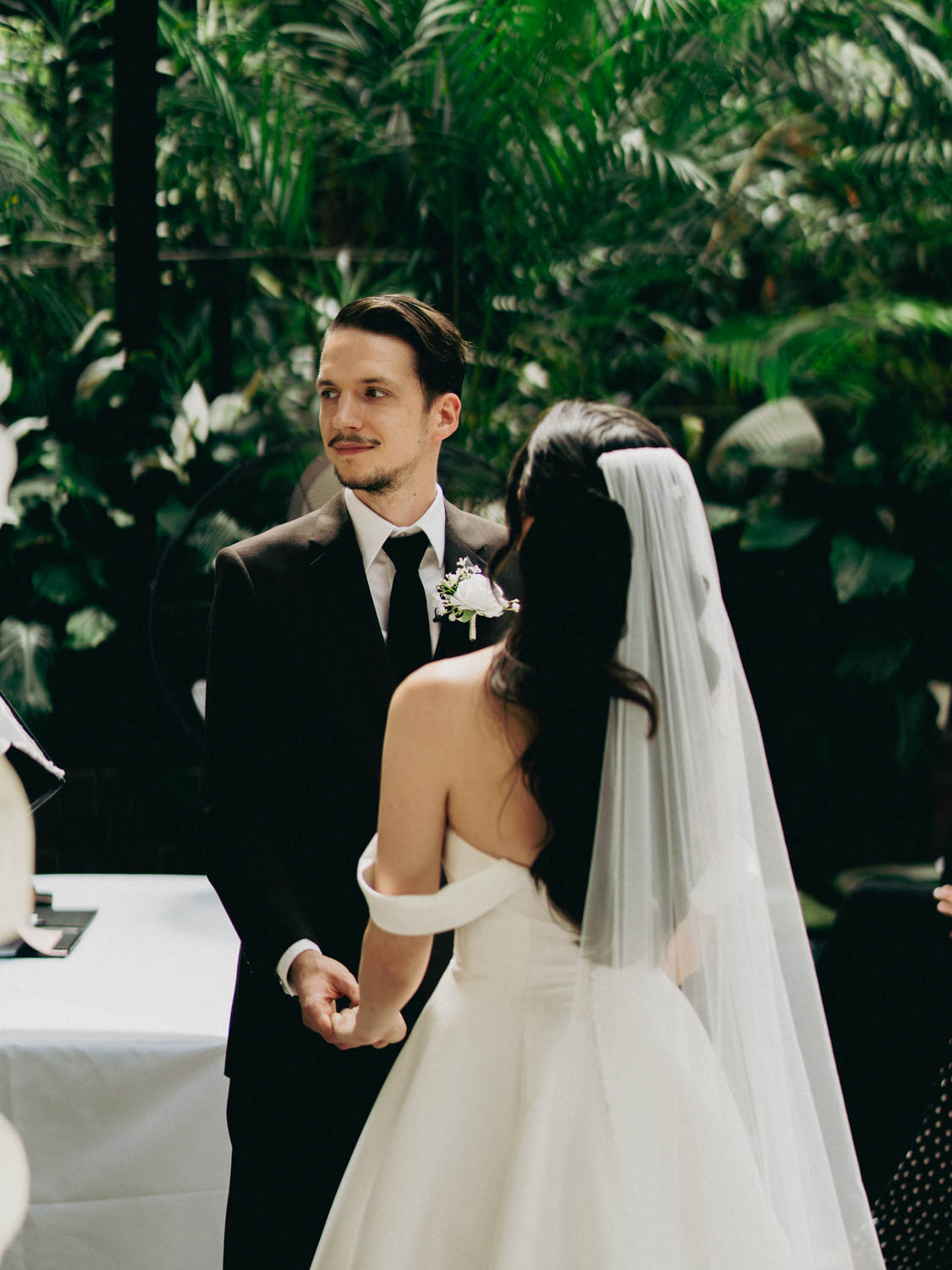 Bride in an off-shoulder white gown and long veil holding hands with groom in a black suit and tie, standing in a lush green indoor setting during their wedding ceremony.
