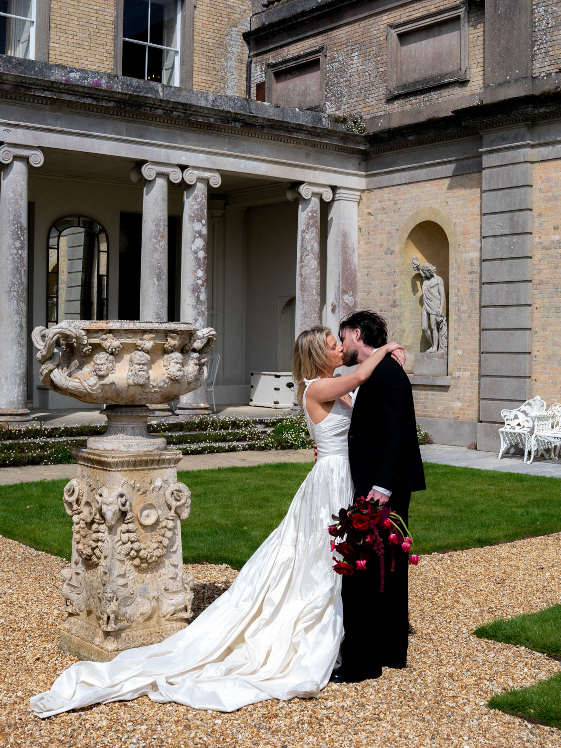 Bride and groom kissing outdoors on a gravel path with green grass and classical architecture in the background; the bride wears a white gown with a long train, and the groom holds a bouquet of dark red flowers.