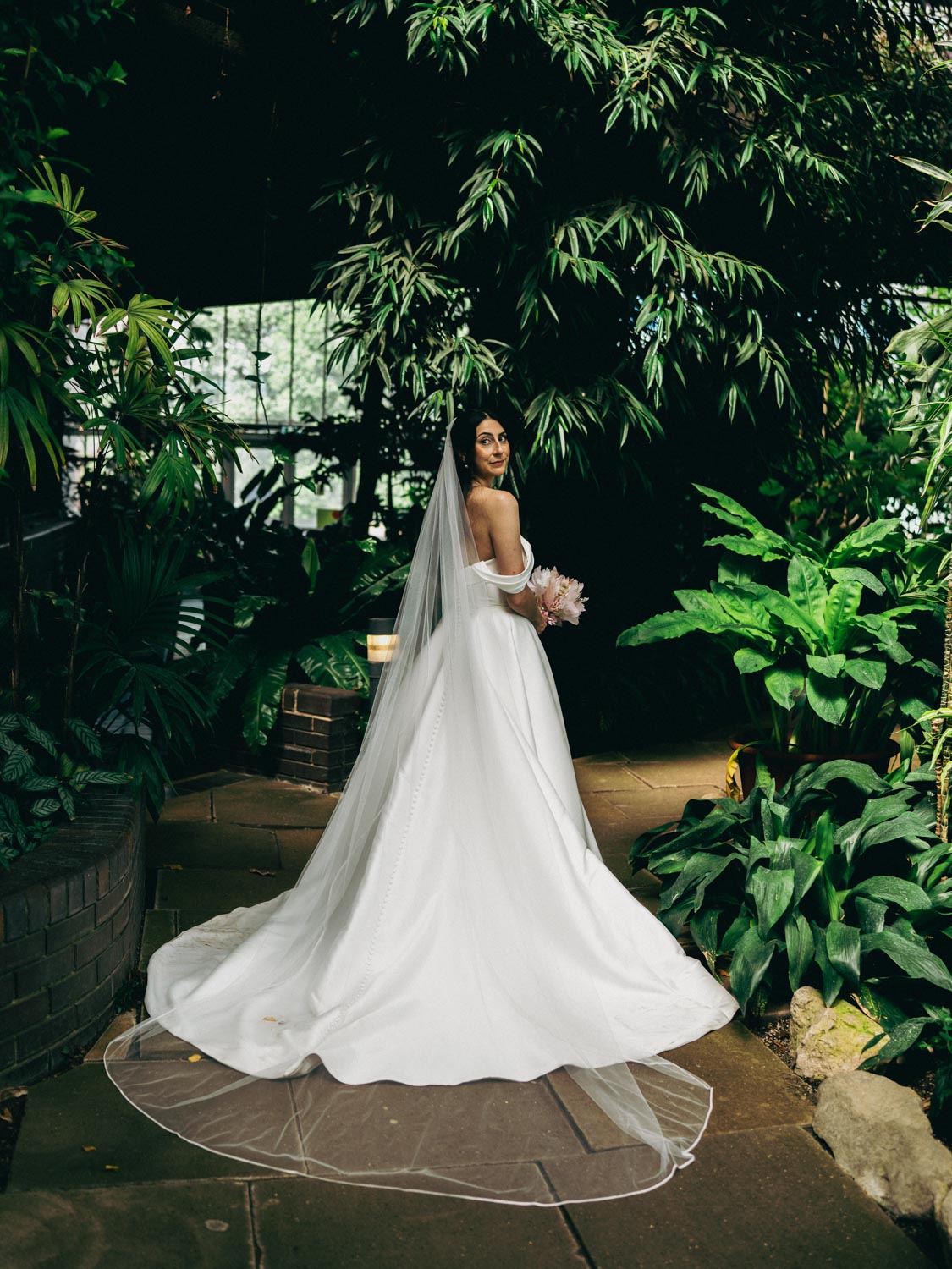 Bride in a white off-the-shoulder wedding dress and long veil holding a pink bouquet, standing on a stone path surrounded by lush green plants indoors.