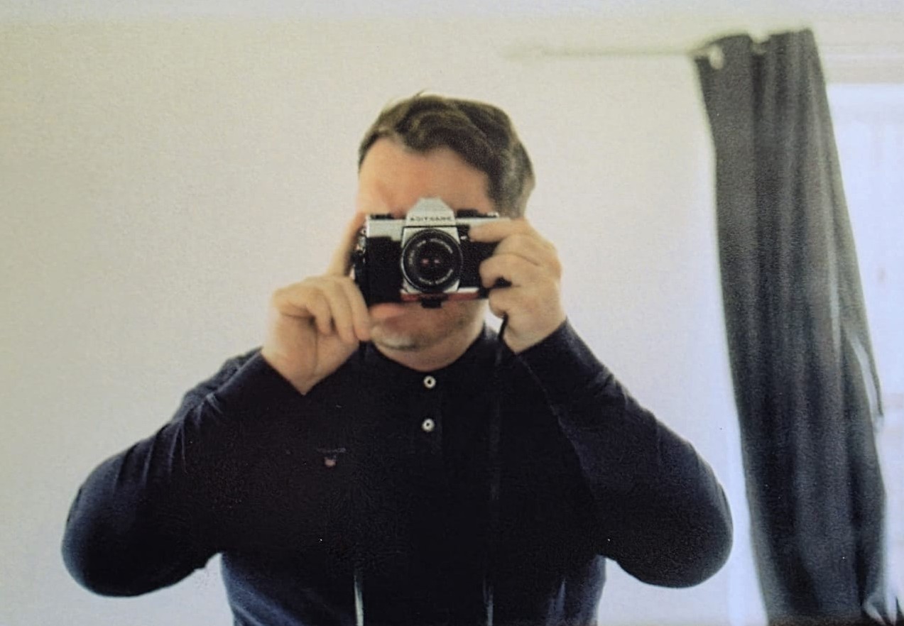 Person wearing a dark long-sleeve shirt taking a photo with a vintage-style camera in front of a plain light-colored wall with a window covered by a dark curtain on the right.