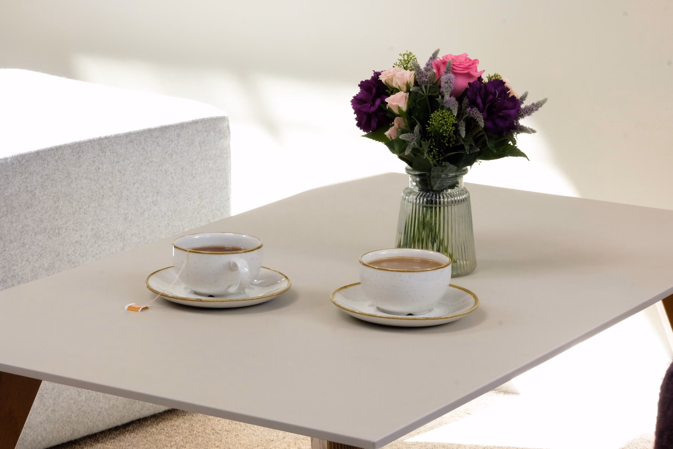 Two white teacups with matching saucers on a light-colored table, alongside a small glass vase holding a bouquet of pink and purple flowers.