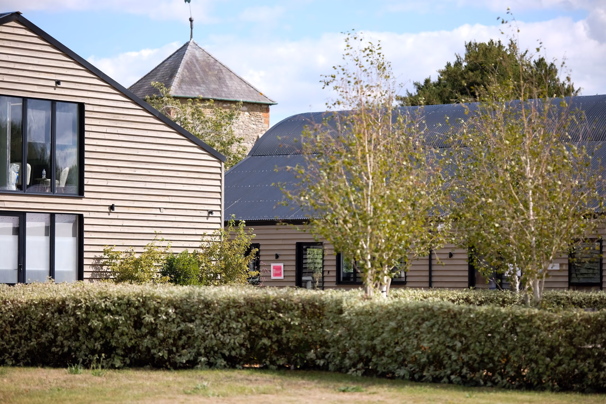 Modern wooden buildings with black framed windows partially obscured by green hedges and small trees under a blue sky with clouds.