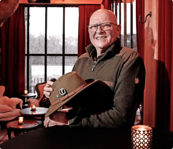 Een oudere man met een bril die binnenshuis glimlacht, een bruine hoed met speldjes vasthoudt en naast brandende kaarsen en een raam met rode gordijnen zit.