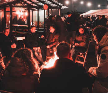 Een groep mensen zit ’s avonds rond een kampvuur, warm gekleed in jassen, met op de achtergrond een verlicht gebouw en sfeervolle lichtslingers buiten.