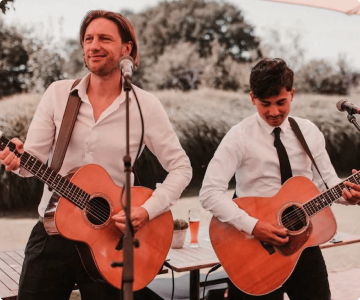 Twee mannen spelen gitaar en zingen buiten bij een microfoon, met een tafel en glazen op de achtergrond.