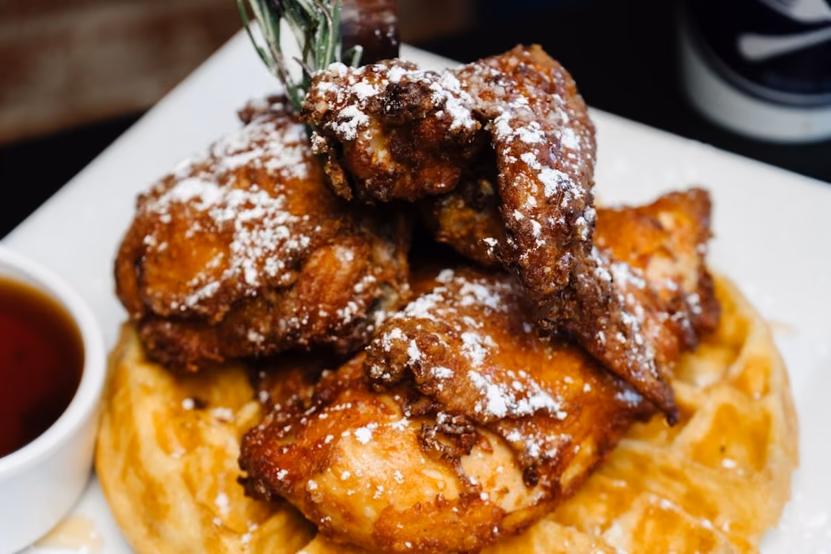 Close-up of crispy fried chicken pieces dusted with powdered sugar, served on a golden waffle with syrup on the side.