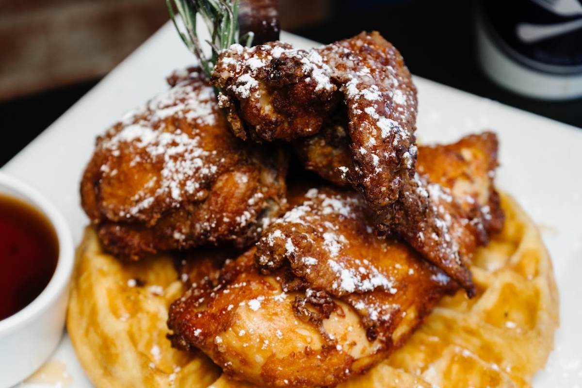 Close-up of crispy fried chicken pieces dusted with powdered sugar, served on a golden waffle with syrup on the side.