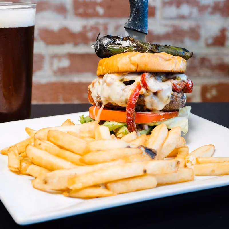 Cheeseburger topped with melted cheese, bacon, lettuce, tomato, and a grilled jalapeño spear held by a knife, served with French fries on a white plate and a glass of dark beer in the background against a brick wall.