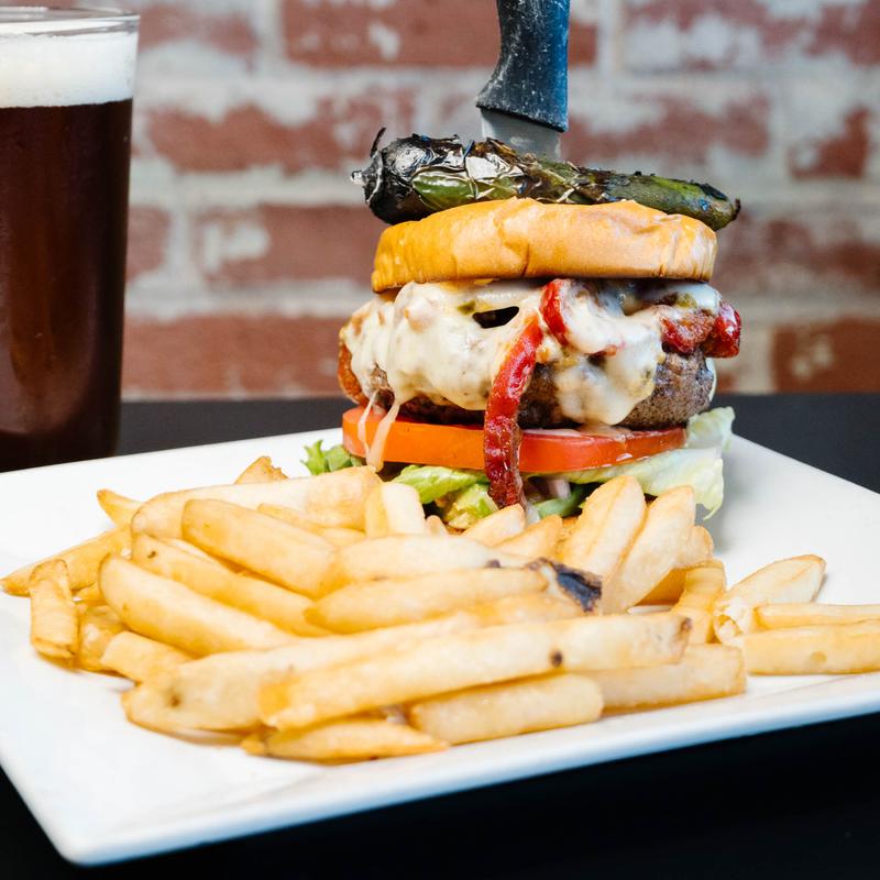 Cheeseburger topped with melted cheese, bacon, lettuce, tomato, and a grilled jalapeño spear held by a knife, served with French fries on a white plate and a glass of dark beer in the background against a brick wall.
