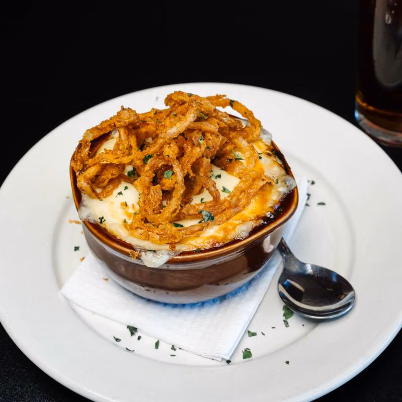 A small brown ceramic bowl filled with baked cheesy dish topped with crispy fried onion strings, garnished with chopped herbs, placed on a white plate with a folded napkin and a spoon beside it.