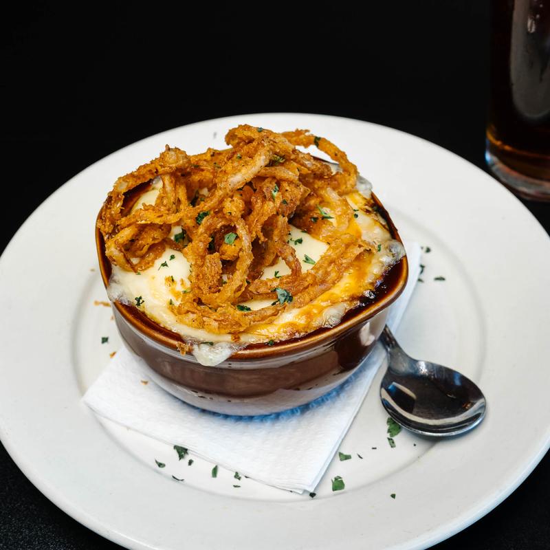 A small brown ceramic bowl filled with baked cheesy dish topped with crispy fried onion strings, garnished with chopped herbs, placed on a white plate with a folded napkin and a spoon beside it.
