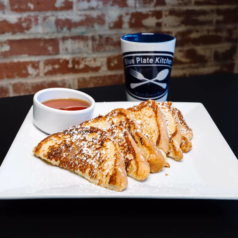 Sliced French toast dusted with powdered sugar on a white plate, served with a small container of syrup and a coffee mug with Blue Plate Kitchen logo against a brick wall background.