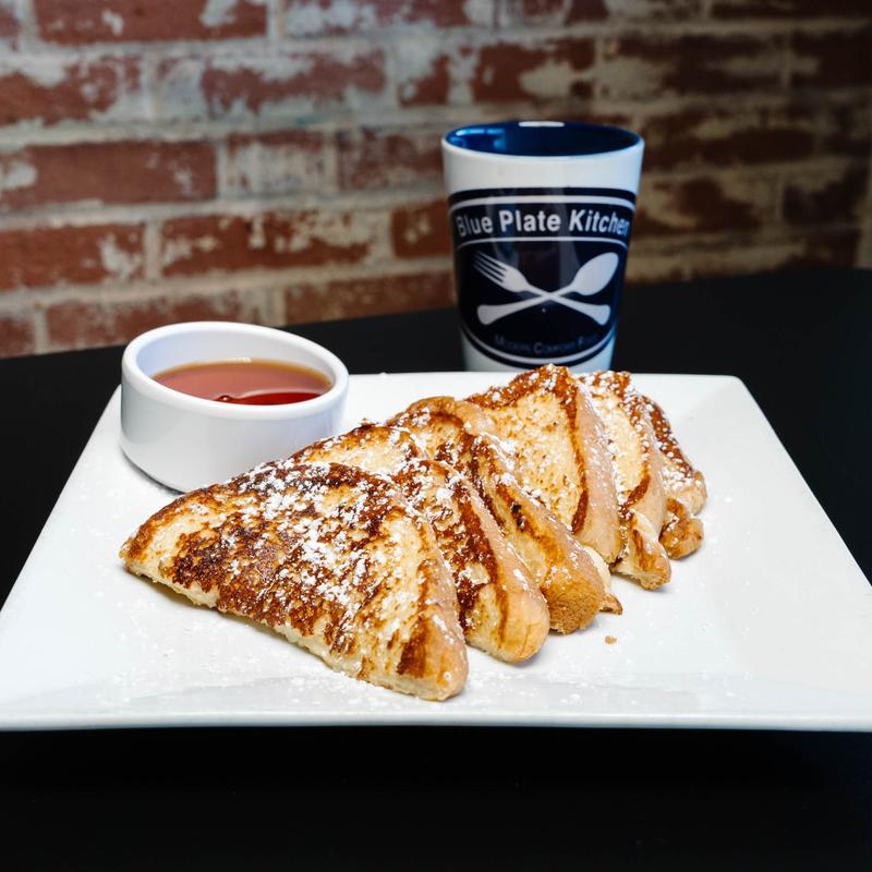 Sliced French toast dusted with powdered sugar on a white plate, served with a small container of syrup and a coffee mug with Blue Plate Kitchen logo against a brick wall background.