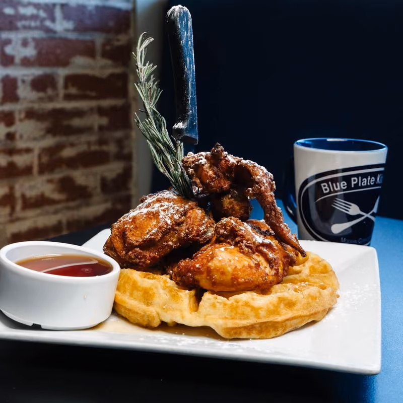 Plate of fried chicken pieces served on a waffle with powdered sugar, garnished with a sprig of rosemary and a knife stuck vertically in the chicken, accompanied by a small bowl of syrup and a mug labeled 'Blue Plate Kitchen'.