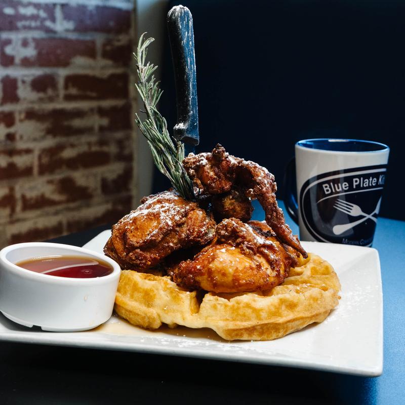 Plate of fried chicken pieces served on a waffle with powdered sugar, garnished with a sprig of rosemary and a knife stuck vertically in the chicken, accompanied by a small bowl of syrup and a mug labeled 'Blue Plate Kitchen'.