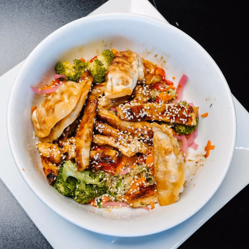 White bowl with grilled tofu strips topped with sesame seeds, pieces of broccoli, pickled red onions, shredded carrots, and three crispy potstickers, placed on a white square plate on a black surface.